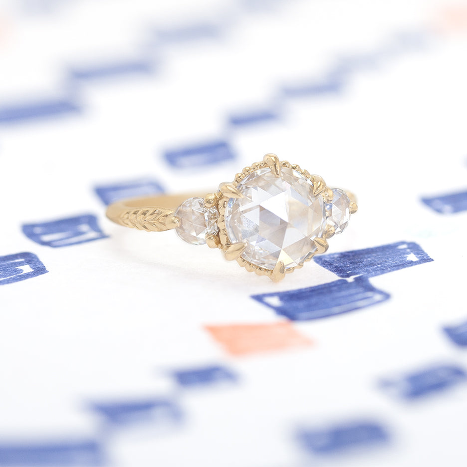 Handmade 3-Stone engagement ring featuring lab grown and natural rose cut diamonds and botanical Evergreen details in 18K yellow gold by Designer Megan Thorne on colored background