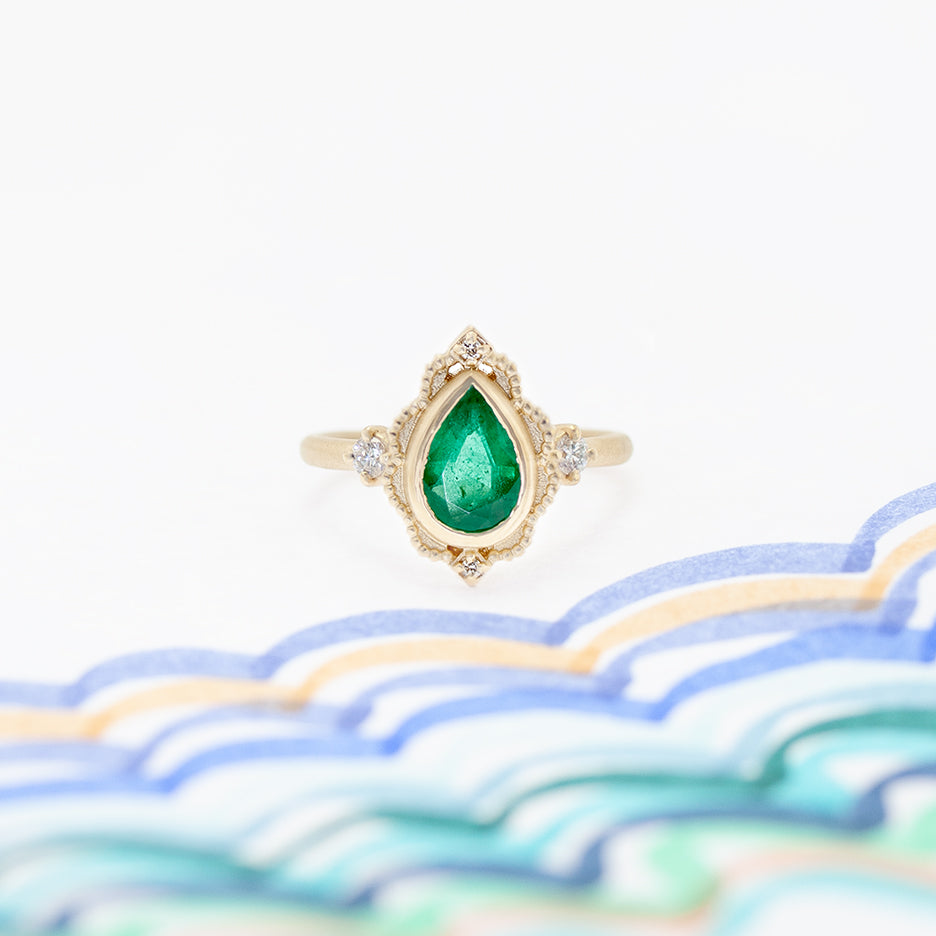 Handmade vintage inspired engagement ring featuring large pear emerald and diamonds with intricated details in 18K yellow gold by designer Megan Thorne on colored background