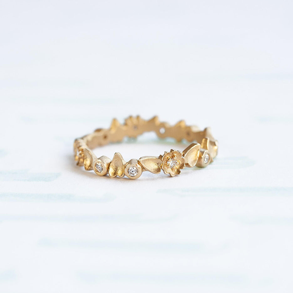Handmade floral eternity wedding band with diamonds and flower and leaf details in 18K yellow gold by Designer Megan Thorne on colored background