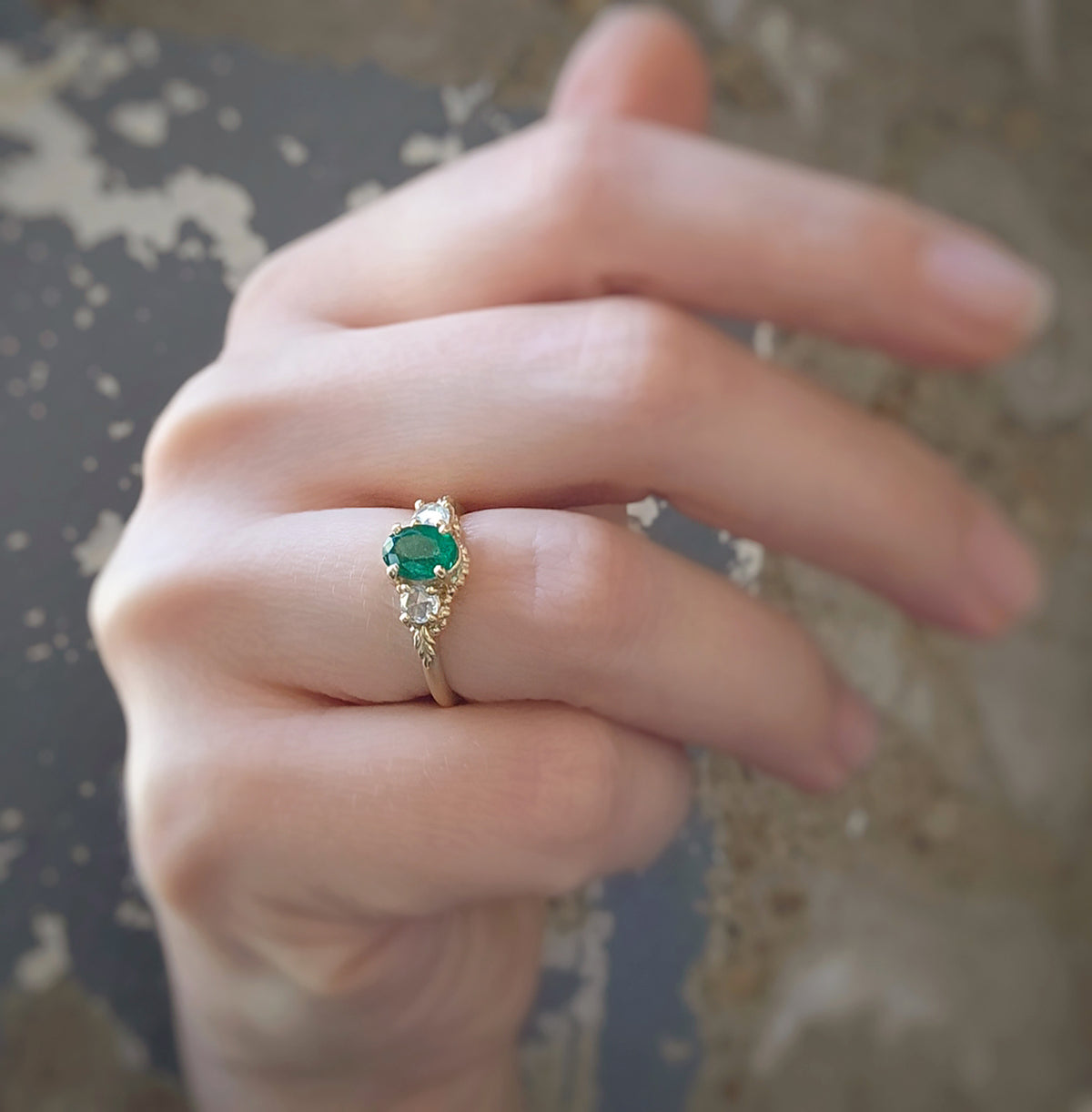A close-up of a hand wearing a gold ring with an oval green emerald center stone and rose cut diamond accent stones.