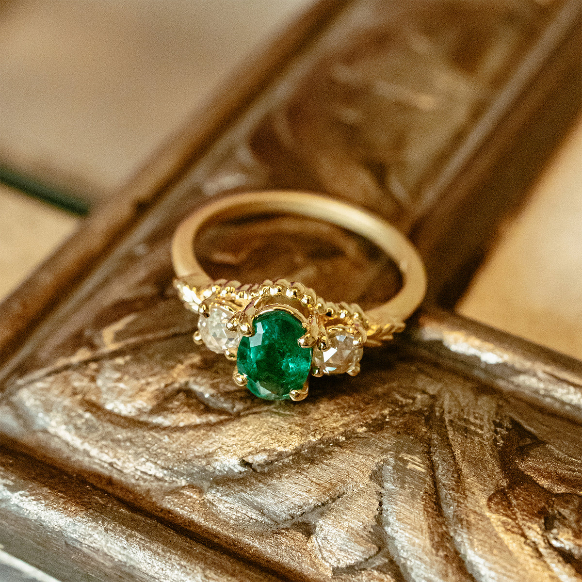 A gold ring with a central oval green emerald gemstone and two small clear rose cut diamond accent stones, resting on an ornate carved wooden surface.