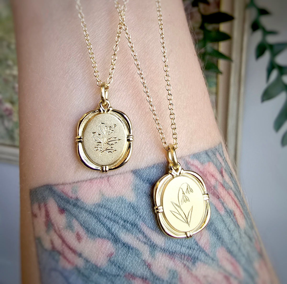 Two gold necklaces with birth flower pendants on a person&#39;s wrist with tattoos.