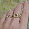 A close-up of a hand wearing two gold rings against a painted background. One ring features a cushion brown diamond in a textured bezel setting, and the other is a delicate band with small clear rose cut diamonds.