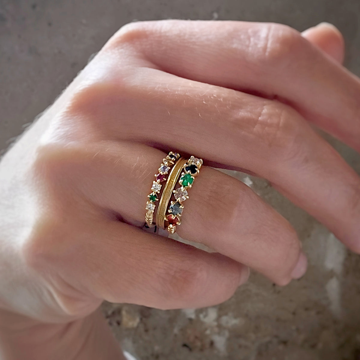 A close-up of a hand wearing a delicate gold ring set with multiple small colorful gemstones, including green, red, and clear stones, against a neutral textured background.