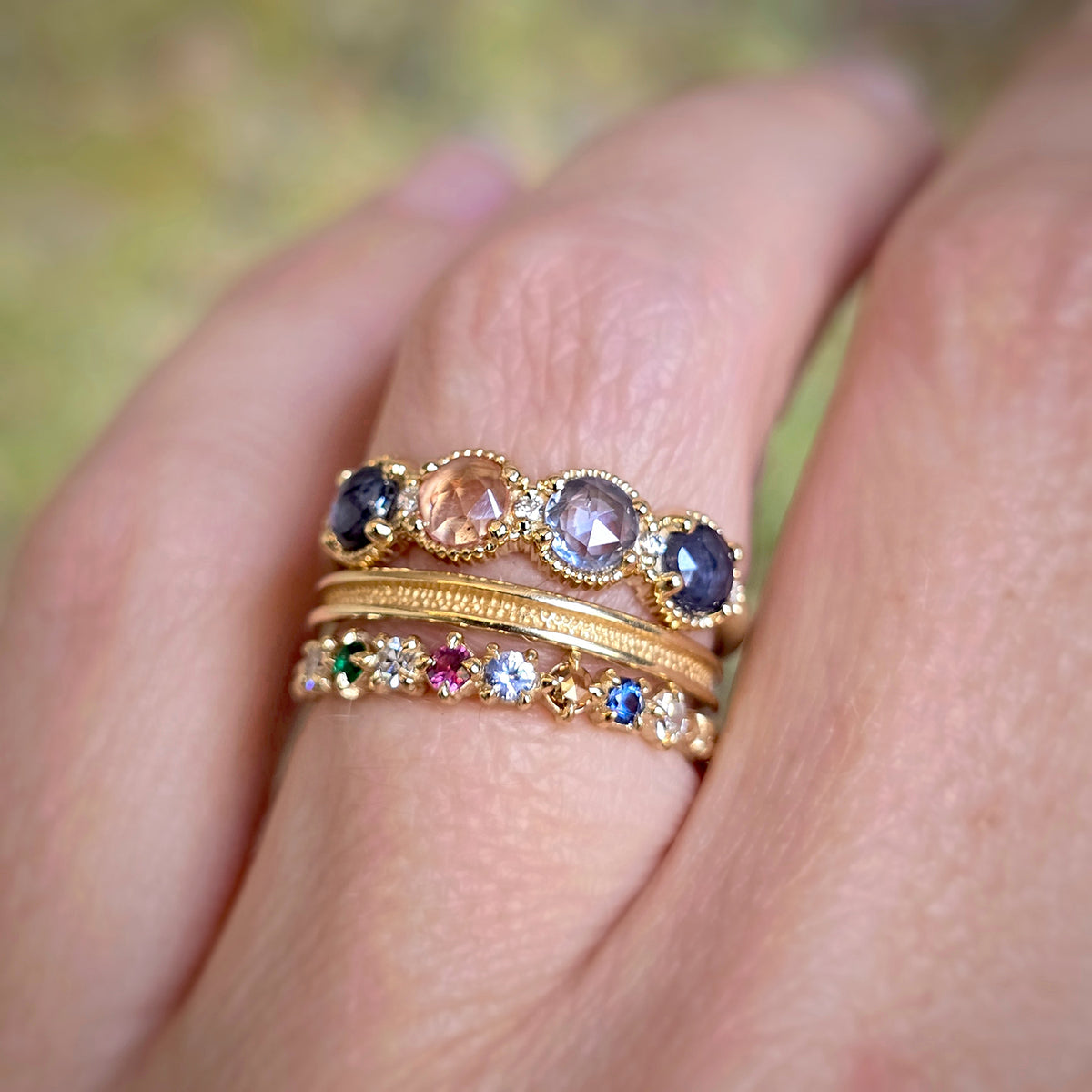 A close-up of a hand wearing three stacked gold rings. The top ring features five oval-cut gemstones in dark blue, peach, and lavender tones.