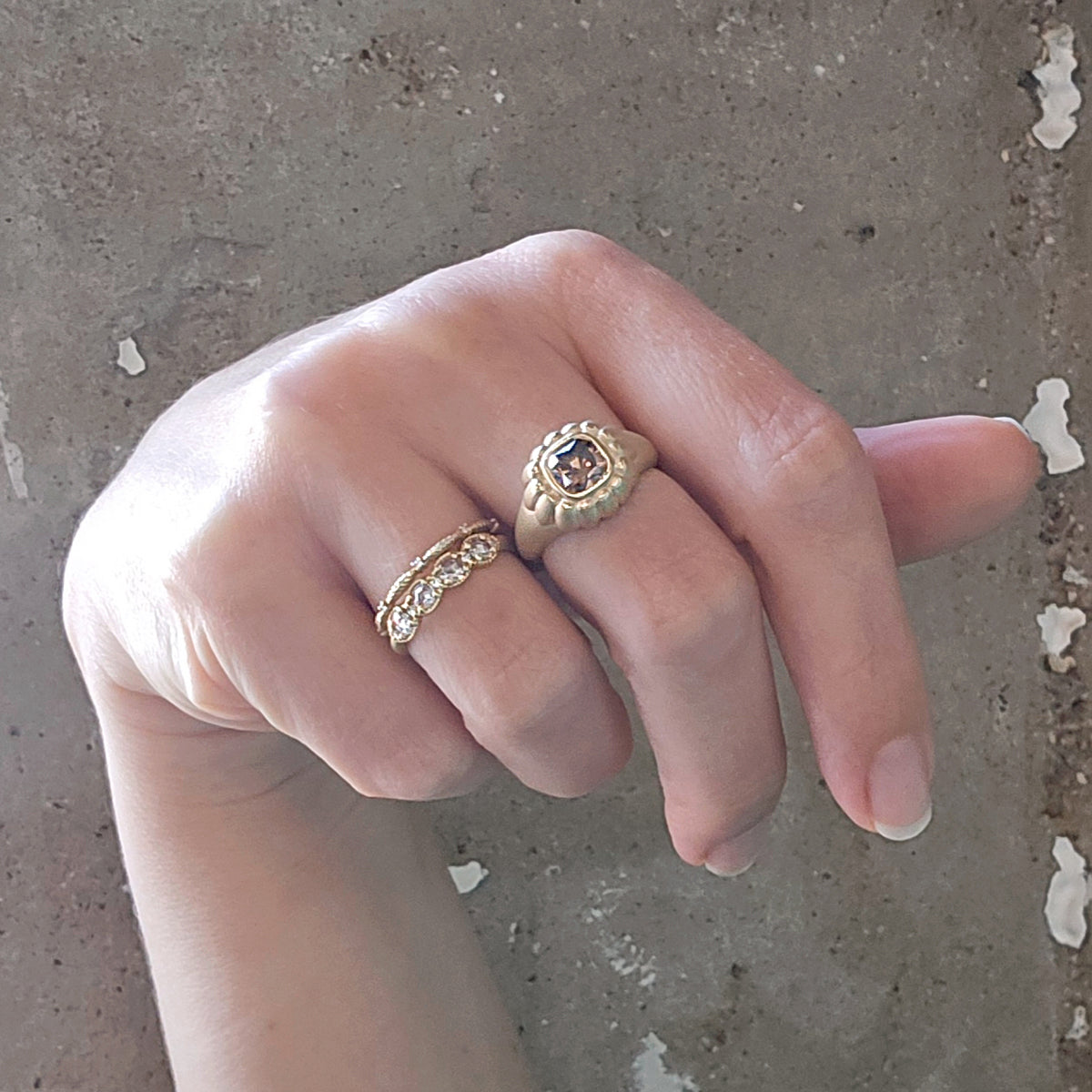 A close-up of a hand wearing two gold rings against a textured concrete background. One ring features a cushion brown diamond in a textured bezel setting, and the other is a delicate band with small clear rose cut diamonds.
