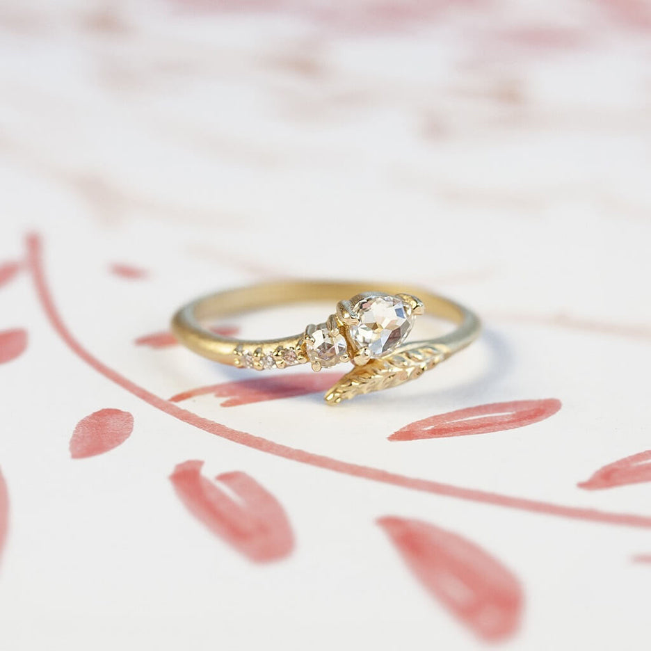 Handmade vintage inspired engagement ring with unexpected Evergreen leaf encircling a pear rose cut diamond and diamond accents in 18K yellow gold by Designer Megan Thorne on colored background