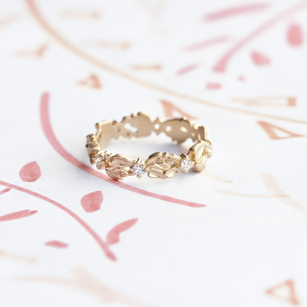Handmade wide eternity wedding band or stacking ring with diamonds and hand carved floral Wood Nymph details in 18K yellow gold by Designer Megan Thorne on colored background